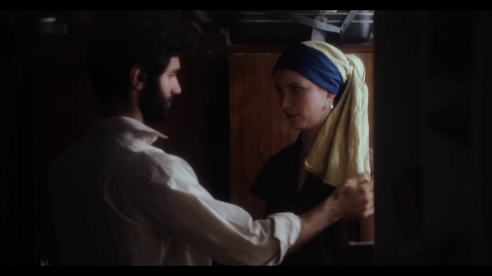 A man and a woman in a dim room, she wears a blue headscarf and a pearl earring (6/8)